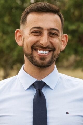 Dr Rami 570x800 - Chiropractor in San Mateo | Quarneri Chiropractic | Accepting New Patients Smiling man with short dark hair and a beard, wearing a light blue dress shirt and navy tie, outdoors with greenery in the background.