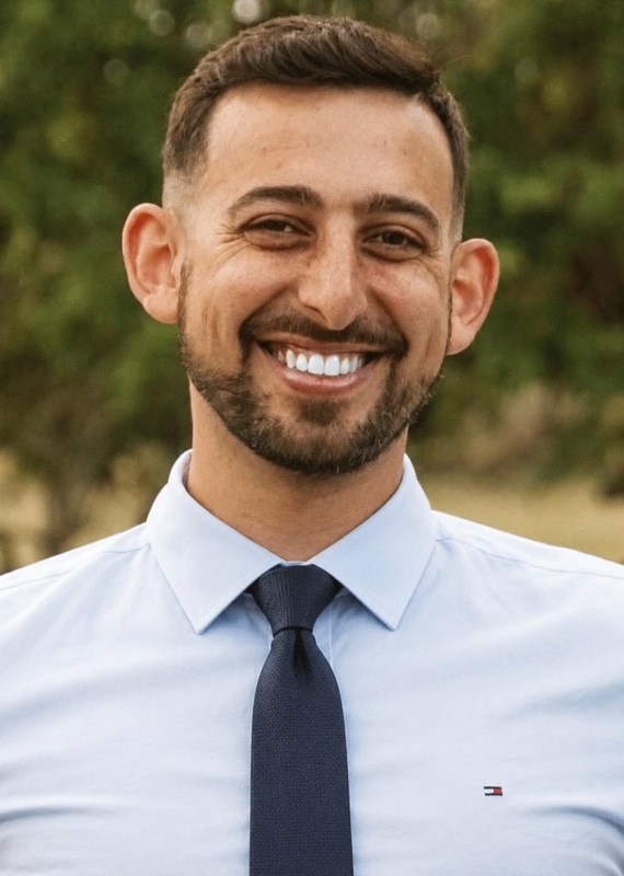 Dr Rami 570x800 - Chiropractor in San Mateo | Quarneri Chiropractic | Accepting New Patients Smiling man with short dark hair and a beard, wearing a light blue dress shirt and navy tie, outdoors with greenery in the background.